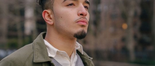 Joven latino con chaqueta verde mira a lo lejos, pensativo, en una calle de la ciudad