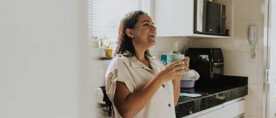 Mujer latina sonriente con una taza de café humeante en la cocina, iluminada por la luz de la mañana