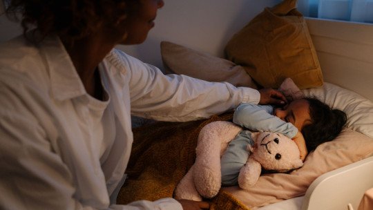 Madre de pelo rizado arropa por la noche a su hija, que duerme abrazada a un osito de peluche bajo una luz cálida