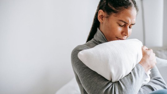 Una mujer joven abraza un cojín con los ojos cerrados, triste, sentada en su habitación