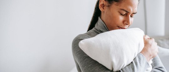 Una mujer joven abraza un cojín con los ojos cerrados, triste, sentada en su habitación