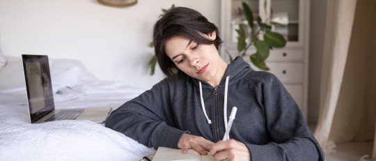 Mujer joven de pelo oscuro escribe en un cuaderno, sentada junto a la cama en una habitación luminosa