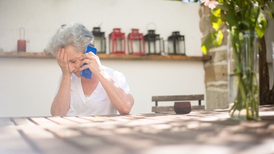 Mujer mayor de pelo canoso, sentada a una mesa al aire libre, habla por teléfono con la mano en la frente