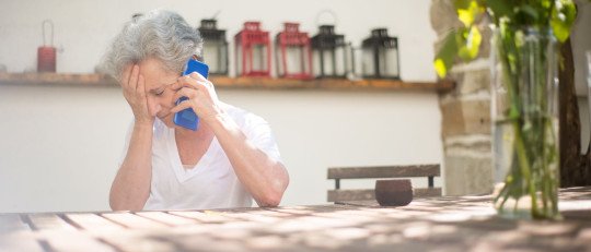 Mujer mayor de pelo canoso, sentada a una mesa al aire libre, habla por teléfono con la mano en la frente
