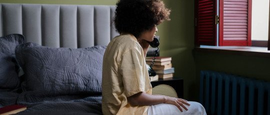 Mujer joven de pelo rizado, sentada en el borde de la cama junto a una ventana, habla por un teléfono fijo