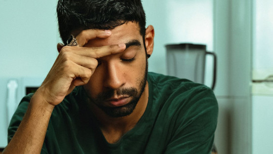 Hombre joven de pelo oscuro y barba, con la mano en la frente y gesto de agotamiento, en una cocina